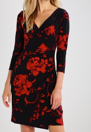 Femme portant une robe noire longueur genou avec des motifs floraux rouges et des manches trois-quarts, debout devant un fond uni.