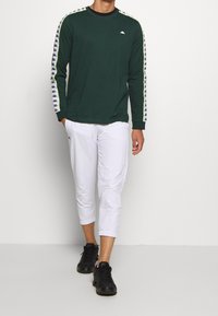 Grön långärmad skjorta med svart krage och Kappa-logotyp, prydd med vita och svarta mönstrade ränder längs ärmarna, i kombination med vita korta byxor.