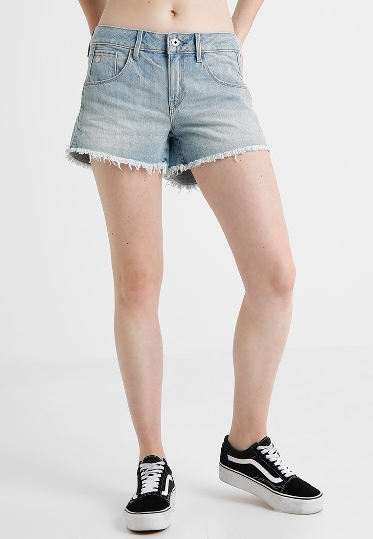 Pantalones cortos de mezclilla azul claro con un dobladillo deshilachado, cinco bolsillos y cierre de botón. Combinados con zapatillas bajas en blanco y negro.