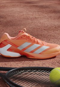 Πορτοκαλί παπούτσι τένις Adidas Adizero σε χωμάτινο γήπεδο δίπλα σε μαύρη ρακέτα τένις και κίτρινη μπάλα τένις.