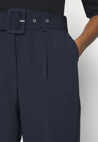 Marinblå högmidjade byxor med ett brett bälte och fyrkantig spänne, veck framtill och sidofickor. Slät tygtextur.