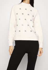 Žena v světle růžovém svetru s opakovaným vzorem "KL" v černé barvě a s černými žebrovanými kalhotami, zobrazená od krku po boky.