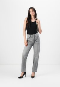 Zwarte bodysuit, grijze high-waisted jeans, elegante zwarte hakken, met een gestructureerde grijze handtas, middellang donker haar, effen witte achtergrond.