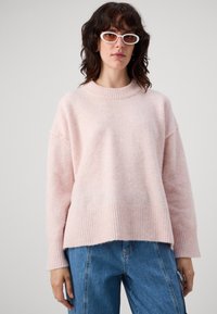 Svijetlo ružičasti, oversized pulover napravljen od mekog, teksturiranog materijala. Ima rebrasti izrez i rub, s spuštenim ramenima i dugim rukavima.