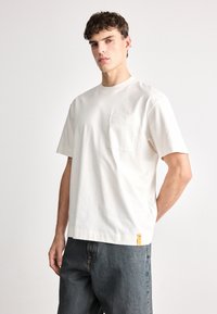 Ανοιχτό μπεζ κοντομάνικο μπλουζάκι φτιαγμένο από βαμβάκι, με τσέπη στο στήθος και μία μικρή κίτρινη ετικέτα στο στρίφωμα. Χαλαρή εφαρμογή με στρογγυλή λαιμόκοψη.