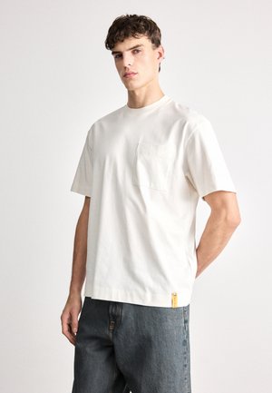 Hellbeige T-Shirt mit kurzen Ärmeln aus Baumwolle, ausgestattet mit einer Brusttasche und einem kleinen gelben Etikett am Saum. Lockerer Schnitt mit Rundhalsausschnitt.