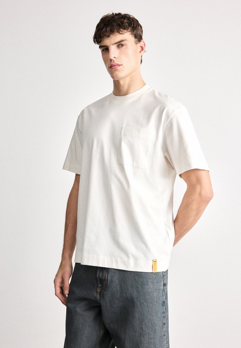 Svijetlo bež majica s kratkim rukavima od pamuka, s džepom na prsima i malom žutom oznakom na rubu. Opusti fit s okruglim izrezom.