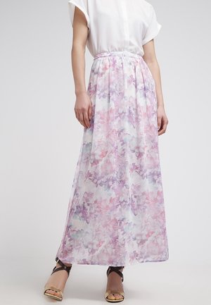 Frau trägt einen langen, fließenden Rock mit einem pastellrosa und violetten Blumenmuster, kombiniert mit einer weißen Kurzarmbluse und offenen High Heels.