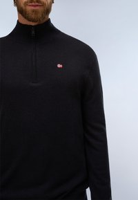 Homme barbu portant un pull noir à quart de zip avec un petit emblème du drapeau norvégien sur la poitrine, sur un fond uni.