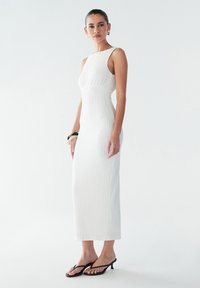 Robe midi blanche à côtes avec une silhouette ajustée, design sans manches et encolure ronde. Le mannequin porte des tongs à talons noirs et des boucles d'oreilles dorées.