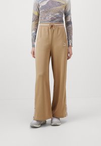Pantalones deportivos PUMA beige con un corte relajado, cintura ajustable con cordón, botones a presión en los laterales y logo bordado en la pierna izquierda. Tela suave y ligera.