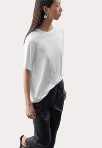 Hvid kortærmet t-shirt med en afslappet pasform, parret med sorte bukser med elastisk talje. Modellen har langt hår og sølvfarvede hoop øreringe.