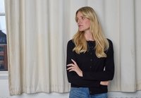 Cardigan nero con bottoni frontali, texture a costine e scollo rotondo, abbinato a jeans blu. Tende di colore chiaro sullo sfondo.