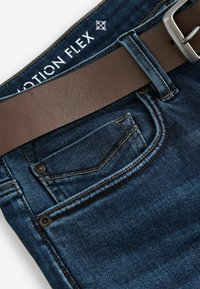 Jeans din denim albastru închis cu o curea din piele maro. Materialul este texturat cu o decolorare subtilă, având un mic buzunar frontal și cusături contrastante.
