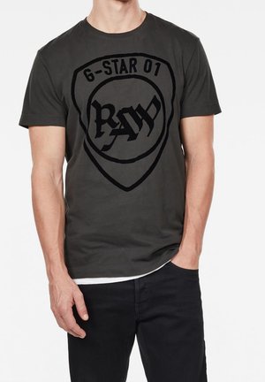 Mężczyzna w ciemnoszarej koszulce z nadrukiem w kształcie tarczy na piersi z tekstem "6-STAR 01" i stylizowanym napisem "RAW", w połączeniu z czarnymi spodniami.