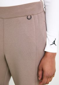 Pantalons de sport marron avec une texture lisse, dotés d'une boucle noire à la taille et d'un logo Jordan noir sur une chemise à manches longues blanche.