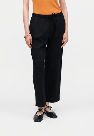 Kvinde iført sorte bukser med vide ben, orange ærmeløs top, sorte snørebåndssko og armbånd på venstre håndled mod en ensfarvet baggrund.