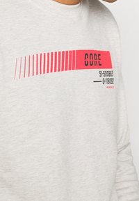 Ljusgrå långärmad skjorta med djärva röda ränder och ordet "CORE" i svart, tillsammans med numeriska detaljer tryckta nedanför.