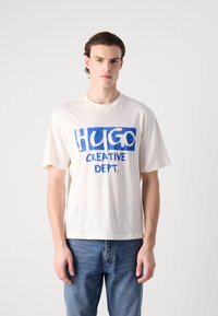 Hvit bomulls T-skjorte med blå grafisk tekst "HUGO CREATIVE DEPT." Korte ermer og en avslappet passform. Kombinert med lyseblå denimjeans.