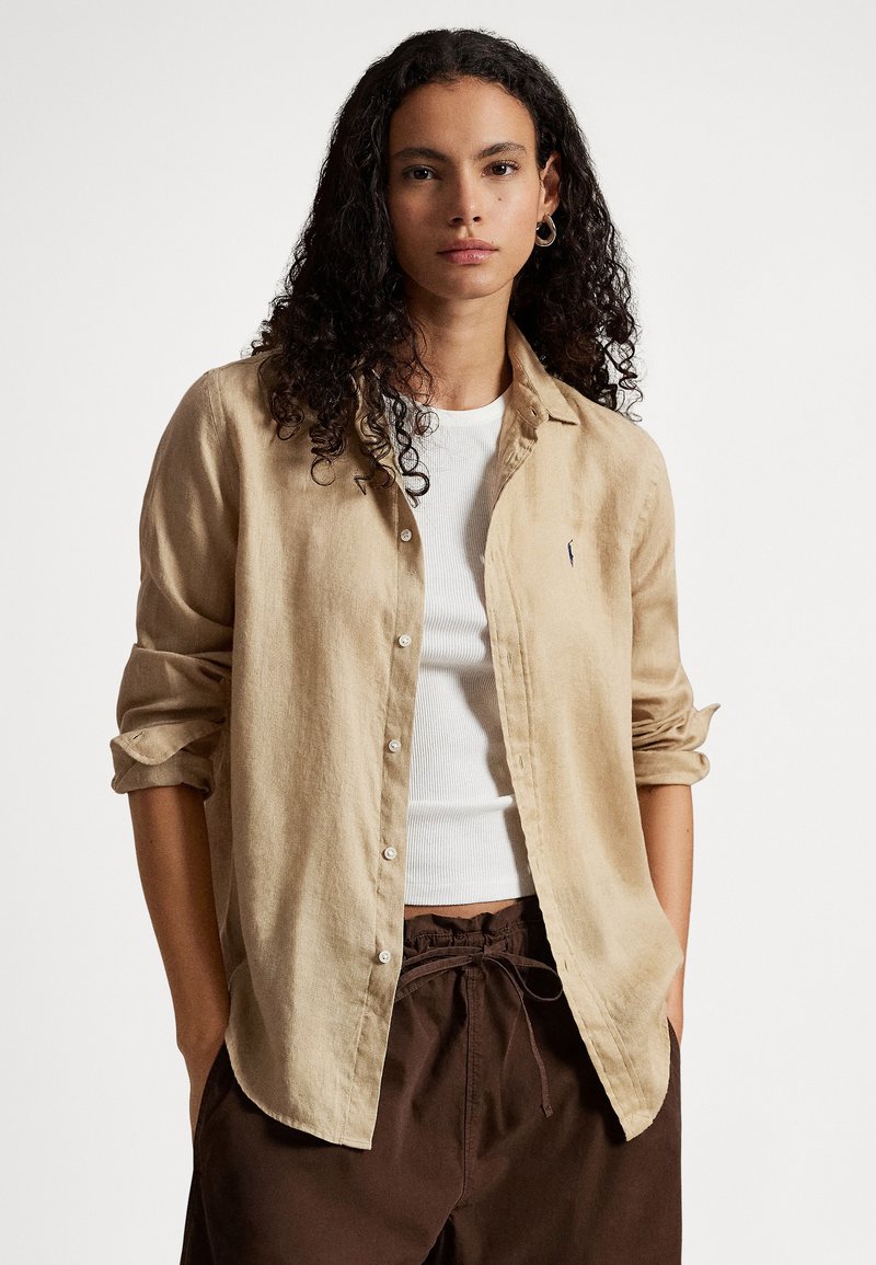 Młoda kobieta o kręconych włosach, ubrana w beżową koszulę na guziki na białym topie oraz ciemnobrązowe spodnie z troczkiem, stojąca z rękami w kieszeniach.