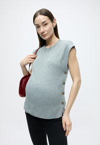 Šedý vrúbkovaný pletený top s krátkymi rukávmi, detailmi na bočných gombíkoch a voľným strihom, skombinovaný s čiernymi nohavicami a malou červenou kabelkou.