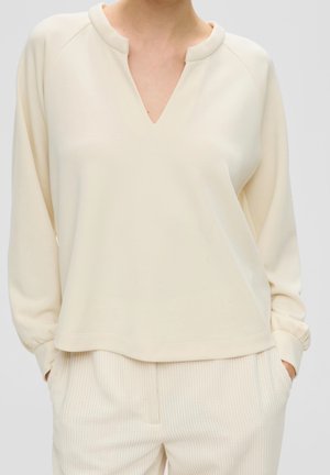 Nainen pukeutuneena kermaisenväriseen pitkähihainen yläosaan, jossa on V-muotoinen pääntie, ja siihen sopiviin kermansävyisiin ribbineulospintaisiin housuihin, kädet taskuissa.