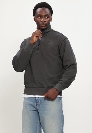Homme portant un pull gris foncé à fermeture éclair au quart et un jean bleu clair, ajustant la fermeture éclair d'une main tout en regardant sur le côté.
