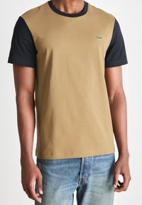 Short-sleeve T-shirt with light brown body and black sleeves; features a small green crocodile logo on the chest; made of cotton.
