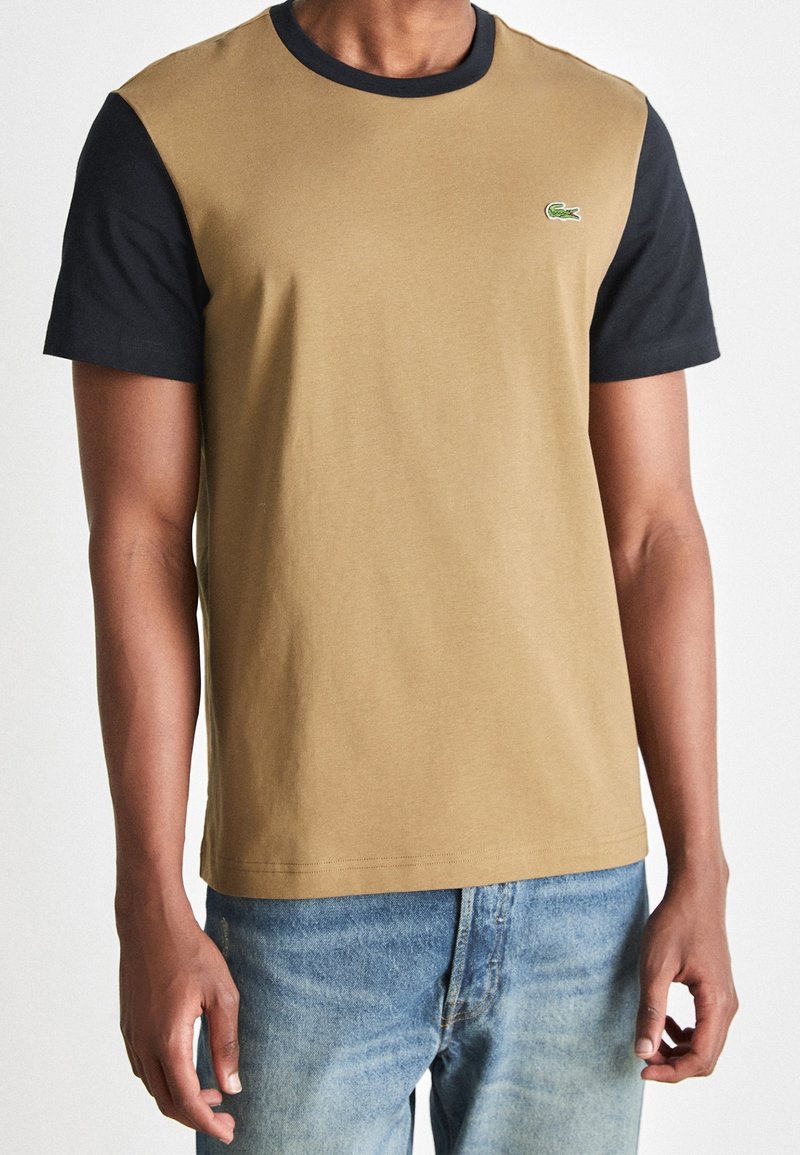 Short-sleeve T-shirt with light brown body and black sleeves; features a small green crocodile logo on the chest; made of cotton.