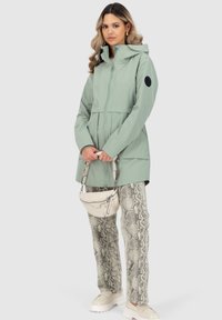 Helle grüne wasserdichte Jacke mit Kapuze, Frontreißverschluss und Taschen. Kombiniert mit einer Hose im Schlangenmuster und einer kleinen cremefarbenen Handtasche.