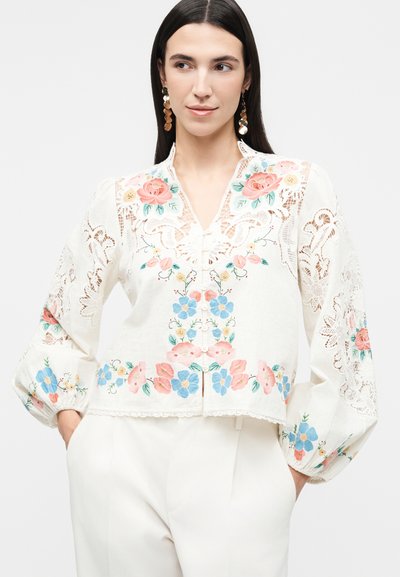 Γυναίκα που φοράει λευκό μπλουζάκι με πολύχρωμη κεντημένη floral διακόσμηση και δαντελωτές ενθέσεις, συνδυασμένο με λευκό παντελόνι και χρυσά σκουλαρίκια δίσκο.