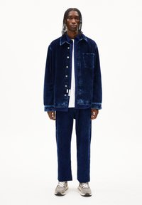Marineblauwe ribfluwelen jas met knoopsluiting en voorzak, gecombineerd met bijpassende ribfluwelen broek. Gekozen voor een witte shirt en grijze sneakers.