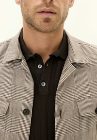 Checkered brown and white jacket with black buttons over a dark brown ribbed polo shirt. The fabric has a soft texture with detailed patterns.
