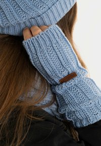 Lichtblauwe gebreide beanie en vingerloze handschoenen hebben een gestructureerd patroon en ribbels aan de randen, met een bruin leren accent op de handschoenen.