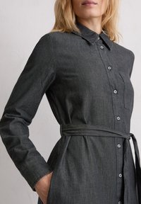 Czarna sukienka koszulowa z dżinsu z długimi rękawami, zapinana na guziki z przodu, z kołnierzykiem, kieszenią na piersi i paskiem w talii. Charakteryzuje się teksturowaną tkaniną oraz subtelnym połyskiem.