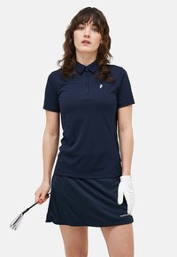Marineblaues Golf-Polo, leichtes Material, kragendes Design, Dreiknopfleiste, weiße Logodetails; kombiniert mit einem passenden Rock und Golfschläger.