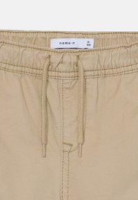 Bézs színű rövidnadrág elasztikus derékrésszel és húzózsinórral. Az anyag sima, enyhén fényes. A derékrészen címke található.