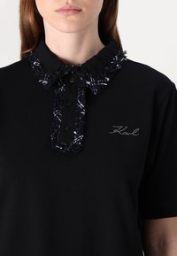 Czarna koszulka polo z teksturowanym, postrzępionym kołnierzykiem w granatowym i białym kolorze. Posiada guziki z przodu oraz haftowane logo na piersi.