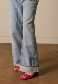 Lichtblauwe wijde jeans met een rafelzoom, gecombineerd met roze suède sandalen met blokhak. Houten vloer en neutrale muur op de achtergrond.