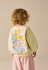 Sárga pulóver színes állat- és virágmintákkal a hátulján, rózsaszín pöttyös nadrággal párosítva. Puha, laza fazon.