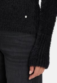 Czarny, puszysty sweter z długimi rękawami, z prążkowanym wzorem i małym srebrnym akcentem przy krawędzi. Zestawiony z ciemnymi jeansami.