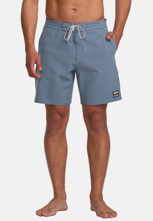 Bañadores de hombre en tela azul claro, con cintura ajustable mediante cordón, bolsillos laterales y un parche de logo colorido en la pierna izquierda.