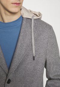 Giacca grigia in misto lana con cappuccio beige e coulisse, indossata sopra una camicia blu. Chiusura con bottoni e tessuto testurizzato.