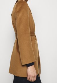 Mujer vestida con un abrigo de lana color camello con cinturón sobre un suéter oscuro y pantalones negros, de pie contra un fondo blanco liso.