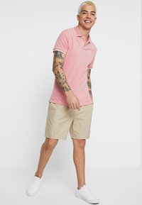 Rød polo-shirt med kontrasterende hvid kant, korte ærmer og logo på venstre bryst. Sammen med beige shorts og hvide sneakers.