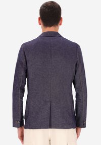 Uomo di spalle che indossa un blazer testurizzato blu scuro e pantaloni color beige chiaro, su uno sfondo bianco uniforme.