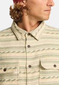 Chemise en flanelle crème et verte avec des motifs géométriques, dotée d'un col étal, d'une fermeture à boutons et de deux poches poitrine avec boutons.