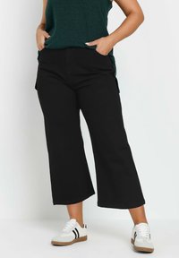 Pantaloni larghi neri realizzati in tessuto testurizzato. Abbinati a una maglietta verde scuro e scarpe bianche con strisce nere. Design casual con tasche.