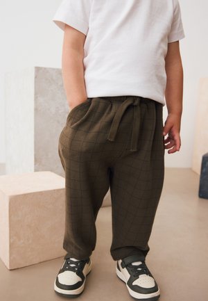 Bambino con maglietta bianca, pantaloni marroni con coulisse e motivo a griglia, e sneakers bianco e nero in piedi con le mani lungo i fianchi.