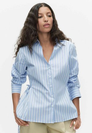 Kvinde med langt krøllet hår iført en lyseblå, hvidstribet skjorte med knapper og rynkede ærmer samt beige bukser.